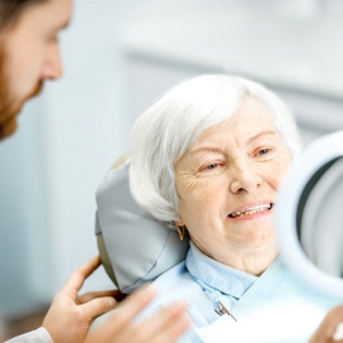 An older woman looking at her smile in the mirror while her dentist stands nearby