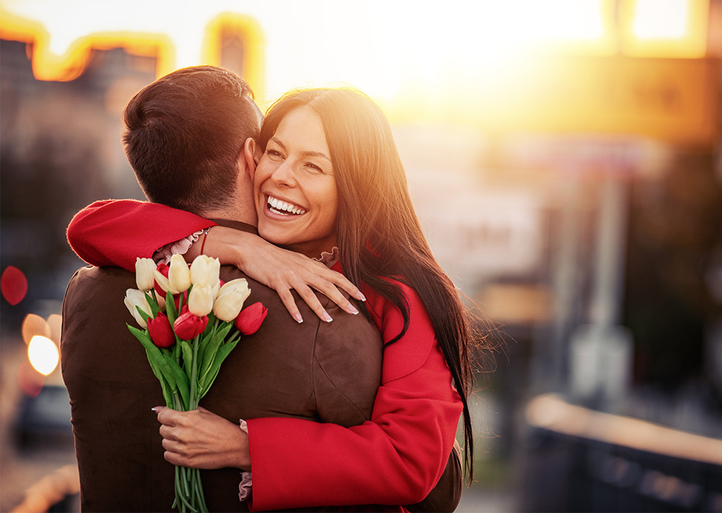 A joyful woman hugs a man while holding a bouquet of tulips, with a sunset in the background.